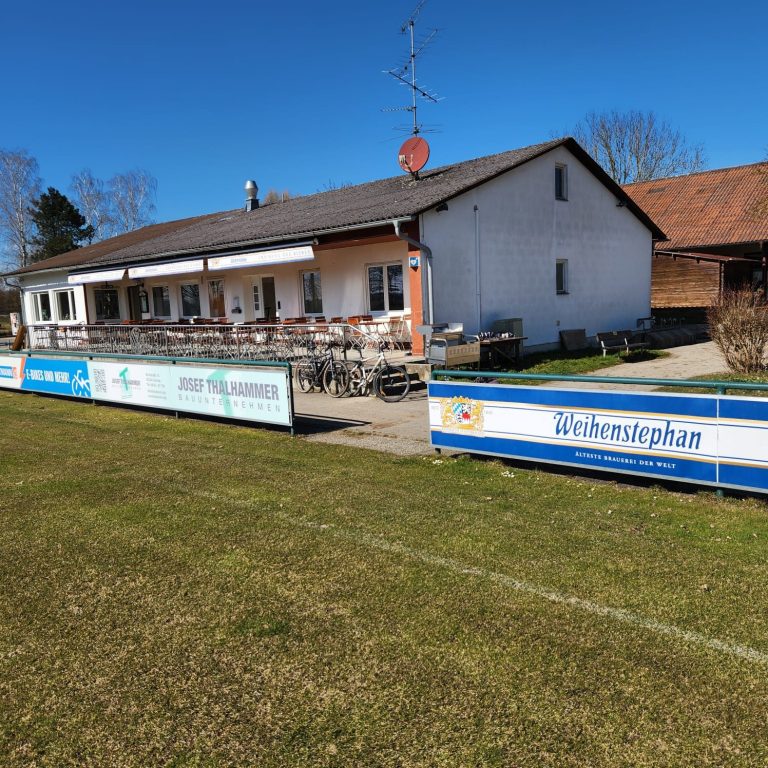 Großes Sportgelände mit einem Vereinsgebäude und einer Terrasse, umgeben von Wiese.